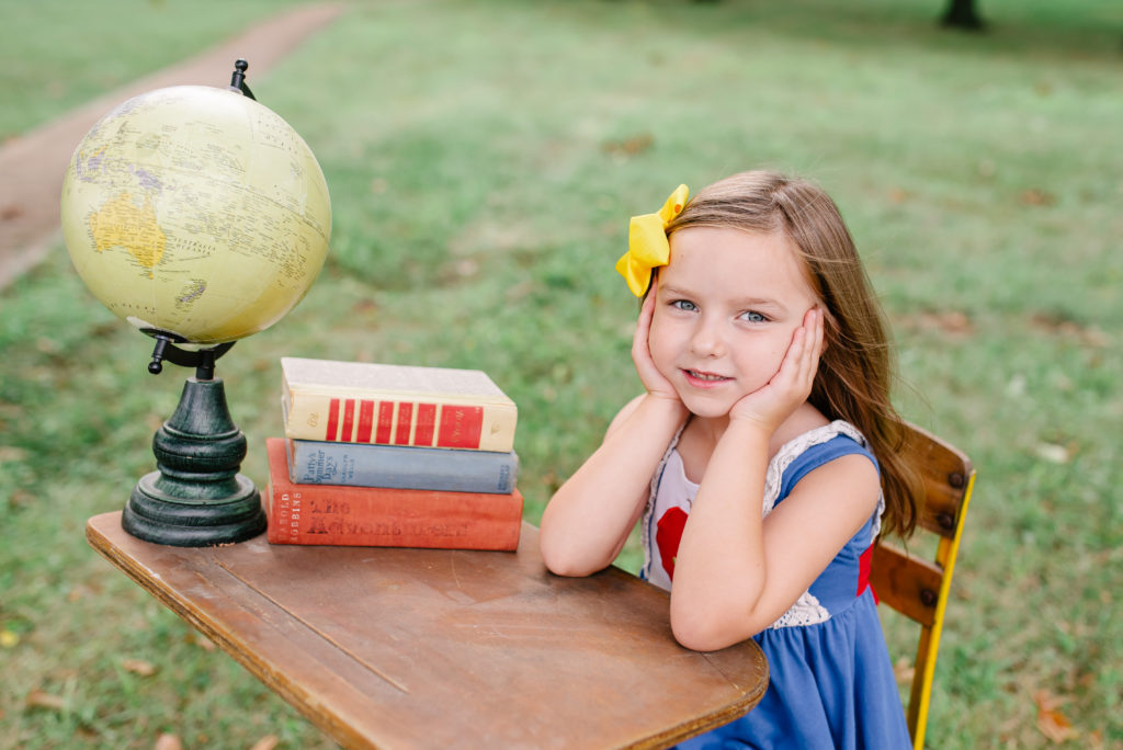 Young girl sits at yellow desk with books for back to school photoshoot.