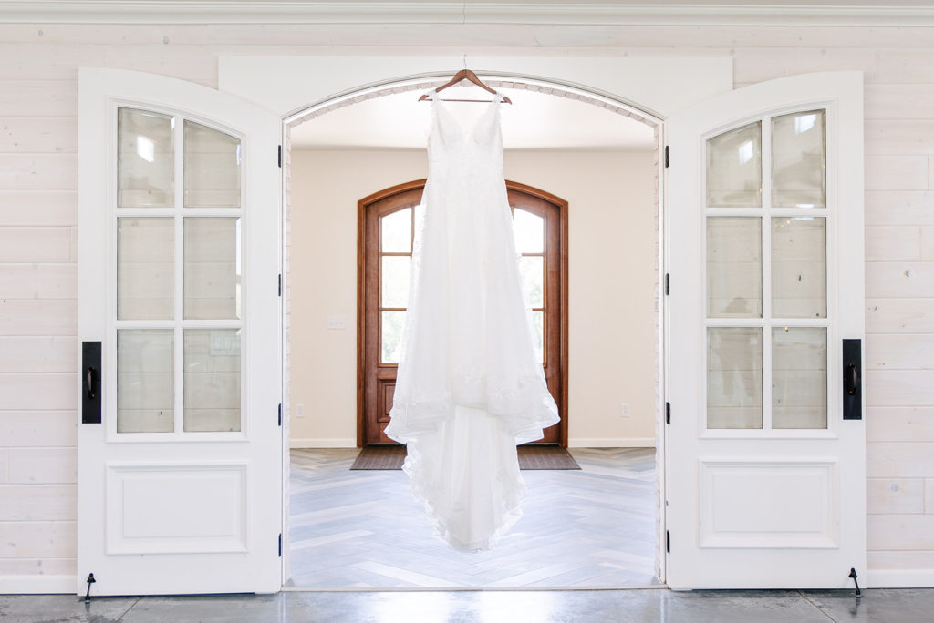 A lace a-line wedding dress hangs from a doorway inside a white barn wedding venue.