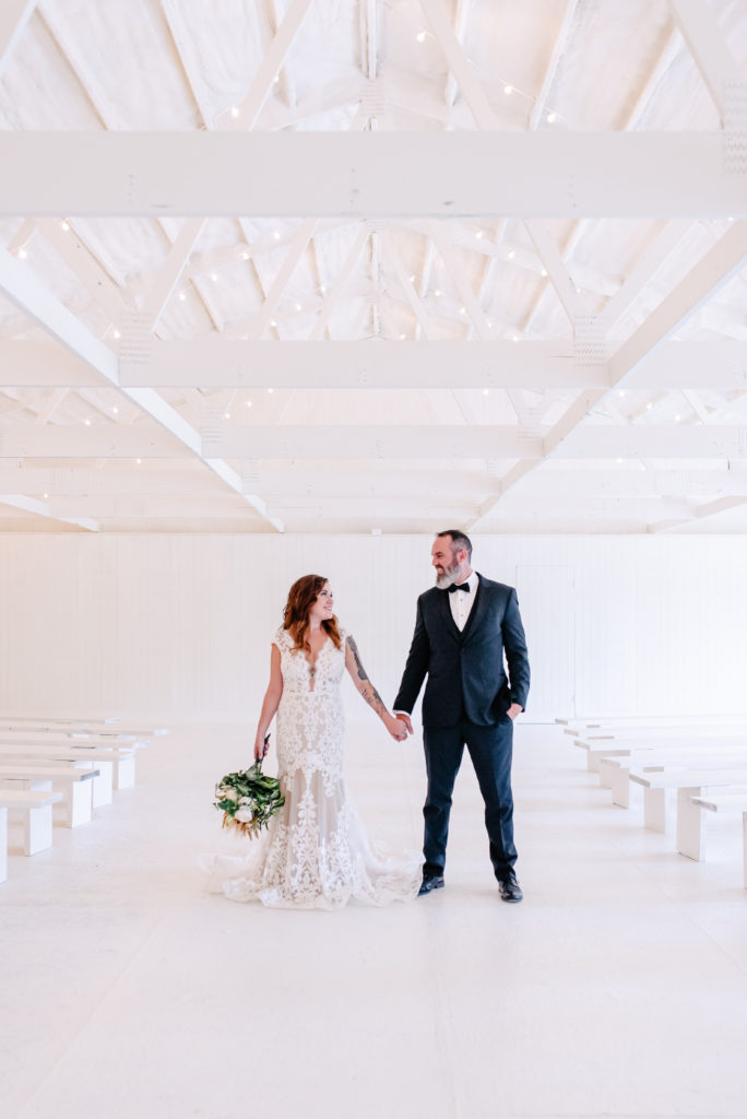 An edgy bride and groom hold hands and pose in the white ceremony room of their wedding venue.
