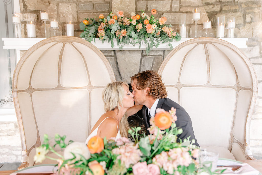 A bride and groom kiss at a pastel and blush sweetheart table adorned with candles.