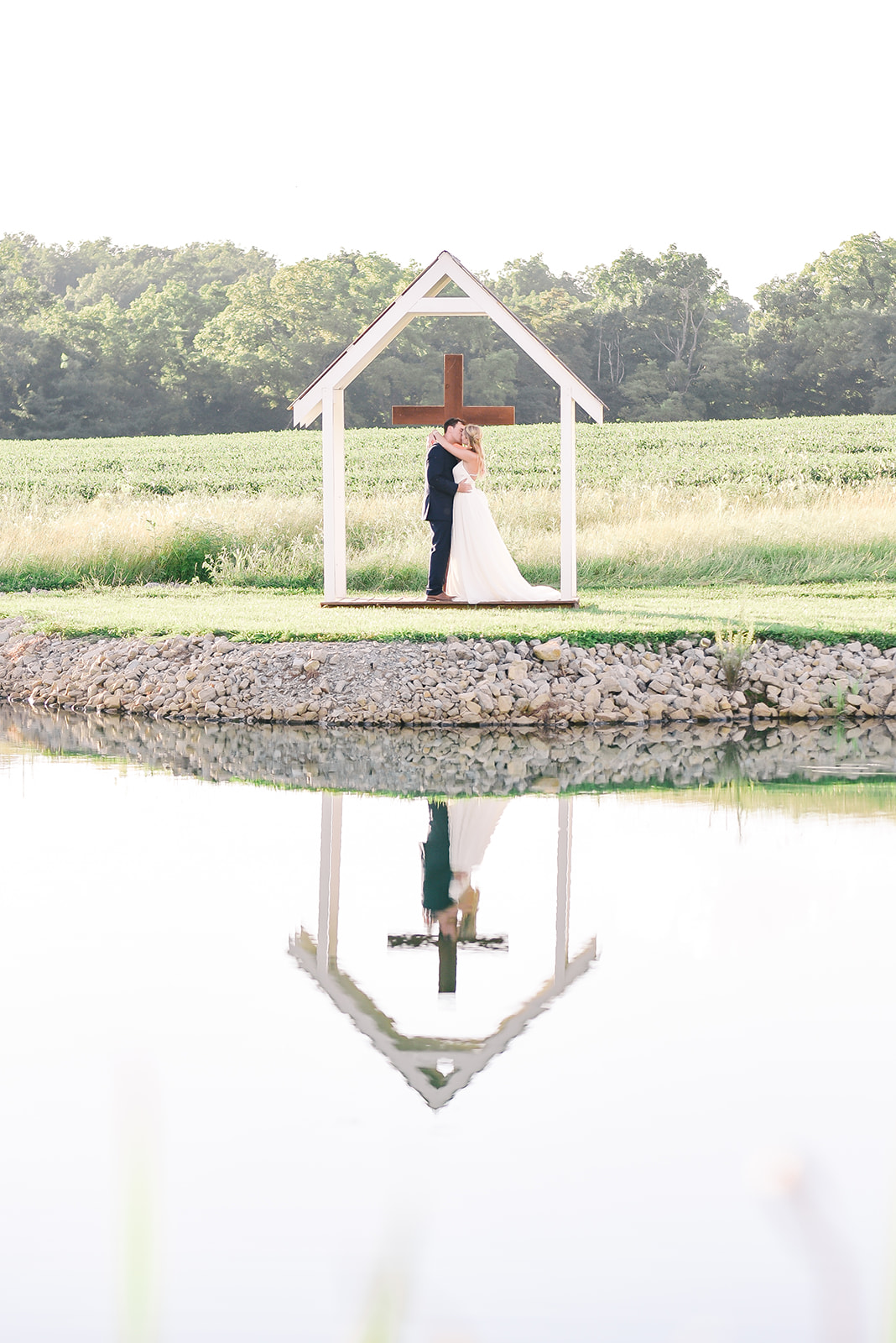 A bride and groom kiss beneath a cross arbor as their reflection glistens on the lake at their wedding venue.