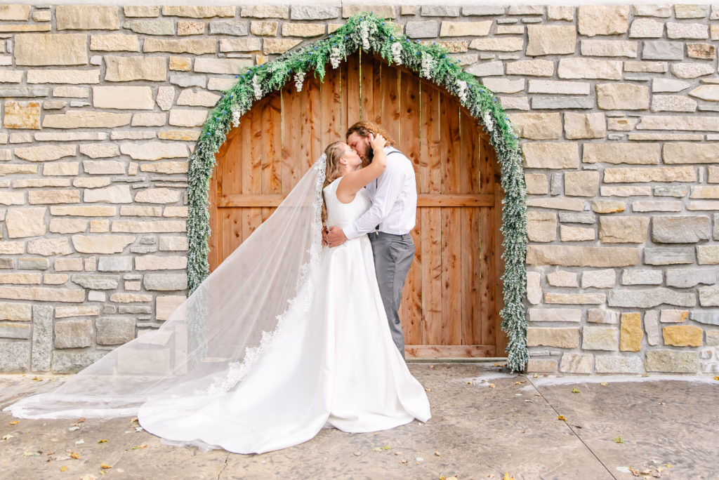 A bride and groom kiss in front of a stone archway on their wedding day.