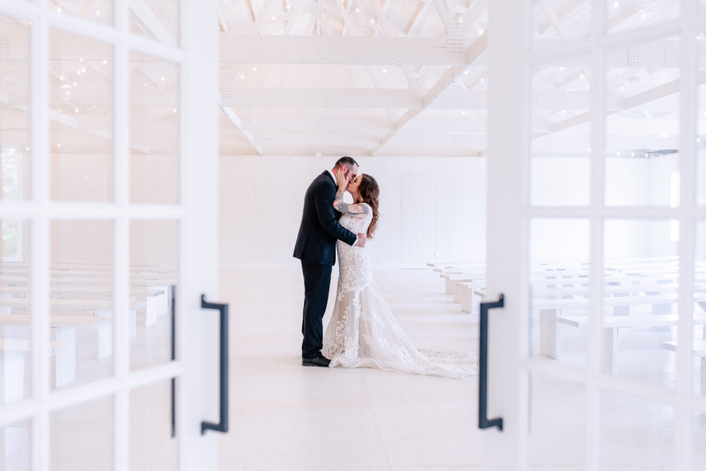 An edgy bride and groom kiss behind closed doors in a white ceremony room on their wedding day.