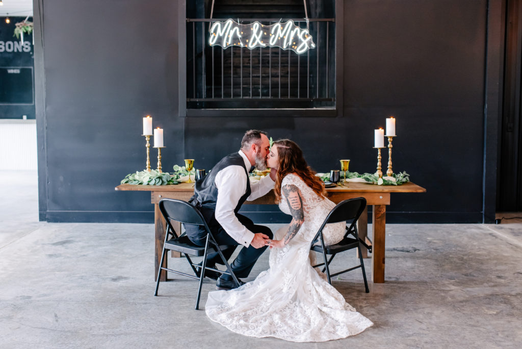 An edgy bride and groom kiss at their sweet heart table with a neon Mr. & Mrs. sign behind them.