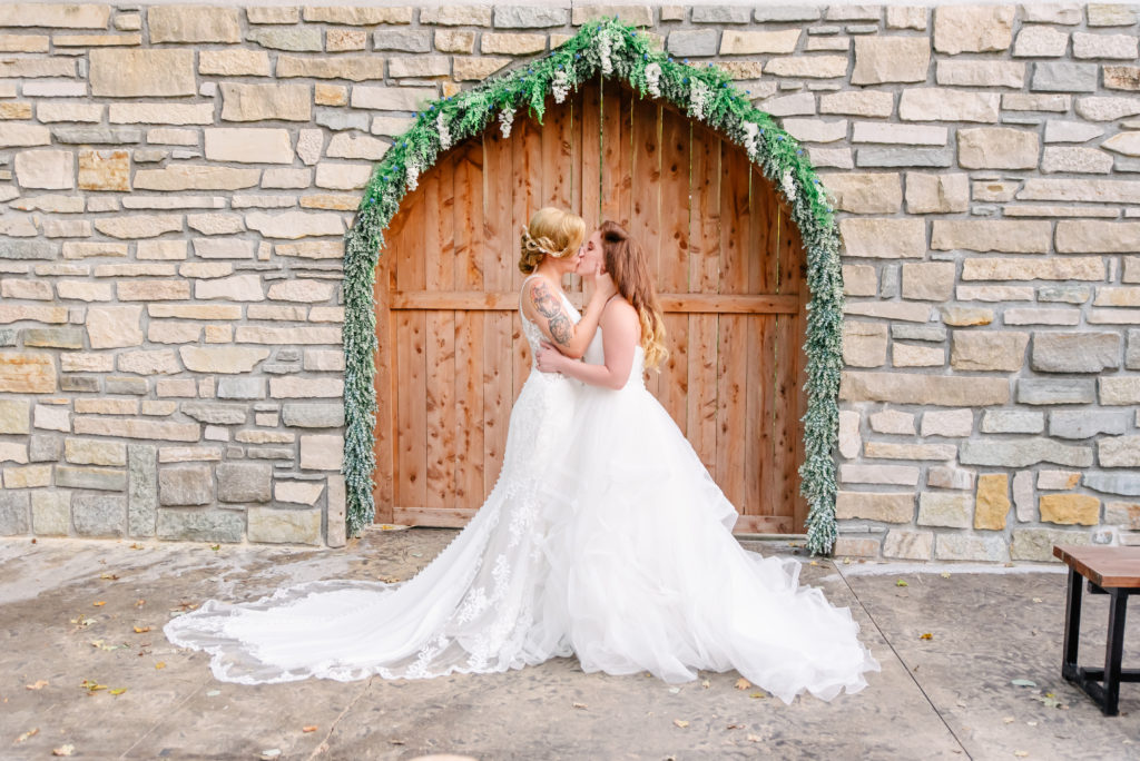 A same sex lesbian couple kiss in front of a stone archway on their wedding day.