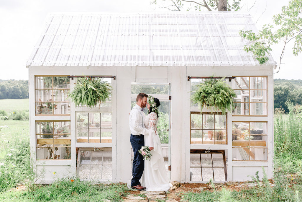 A bride and groom snuggle nose to nose in front of a rustic greenhouse on their wedding day.
